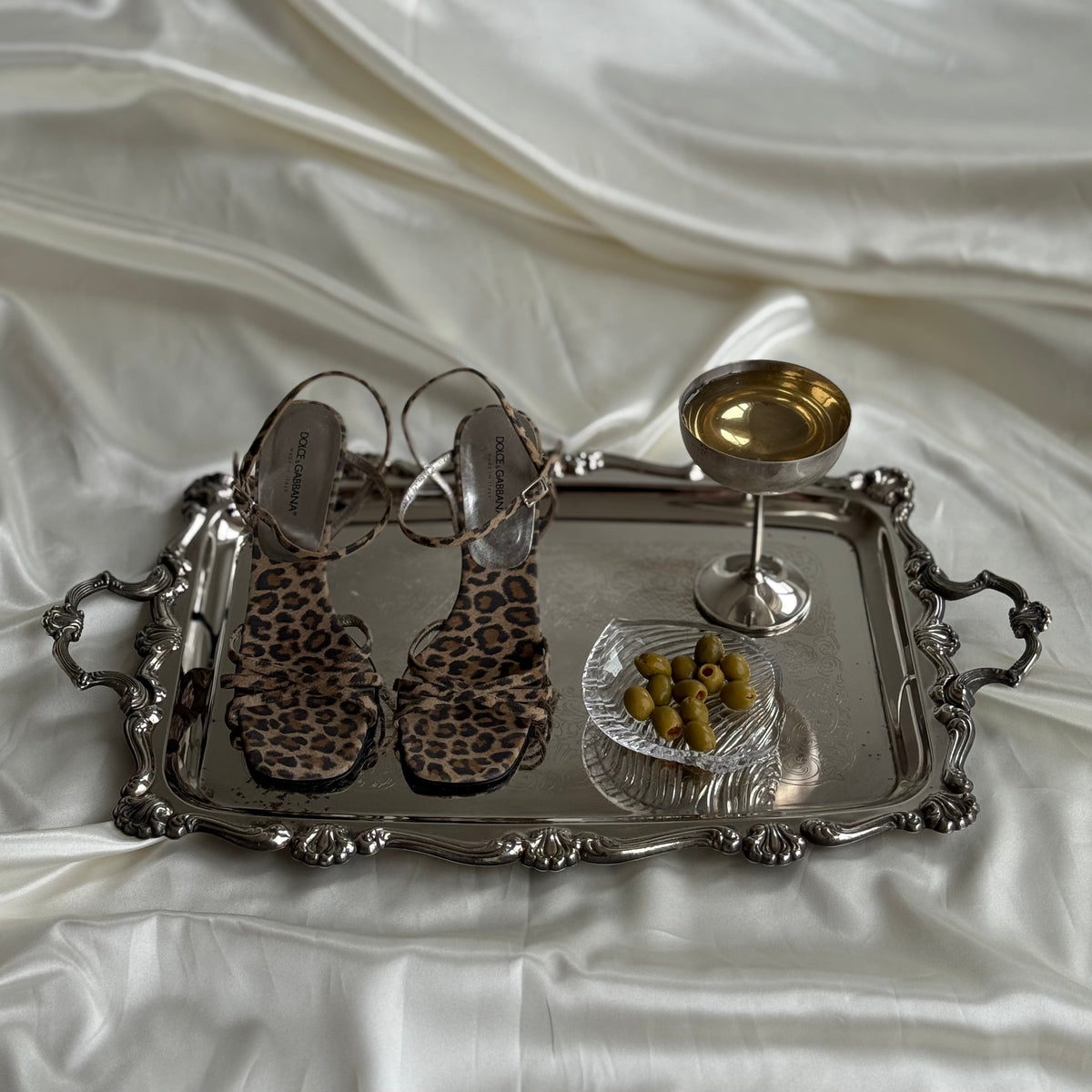 Leopard print flat sandals with strappy heels arranged on an ornate silver tray, alongside a small bowl of olives and a gold goblet, all set on draped white satin fabric.